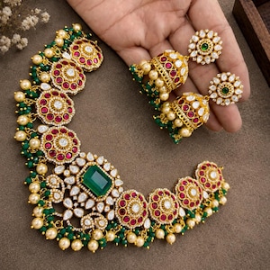 May include: A gold-toned necklace and matching jhumka earring set featuring a central rectangular green stone, circular floral motifs with red stones, white accents, and dangling green and cream-colored beads throughout the jewelry pieces.
