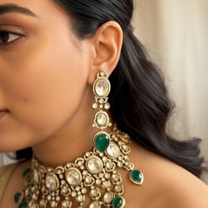 Op de afbeelding: Close-up van een goudkleurige ketting en bijpassende oorbellen. De sieraden hebben heldere, ovaalvormige stenen en levendige groene druppelvormige stenen. De oorbellen zijn lang en bungelen, terwijl de ketting dicht op de nek zit.