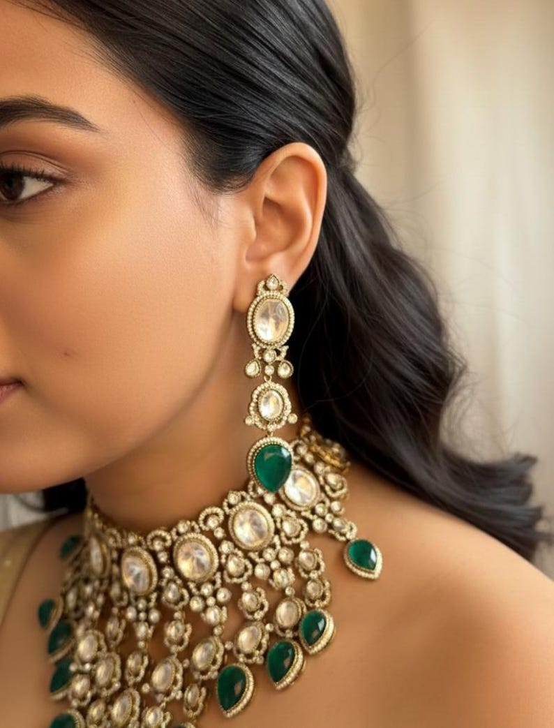 Op de afbeelding: Close-up van een goudkleurige ketting en bijpassende oorbellen. De sieraden hebben heldere, ovaalvormige stenen en levendige groene druppelvormige stenen. De oorbellen zijn lang en bungelen, terwijl de ketting dicht op de nek zit.