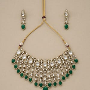 Op de afbeelding: Een goudkleurige ketting- en oorbellenset. De ketting heeft ovale en ronde heldere stenen met druppelvormige groene stenen. De oorbellen weerspiegelen het ontwerp met een enkele groene druppelsteen. De set wordt gepresenteerd op een neutrale achtergrond.