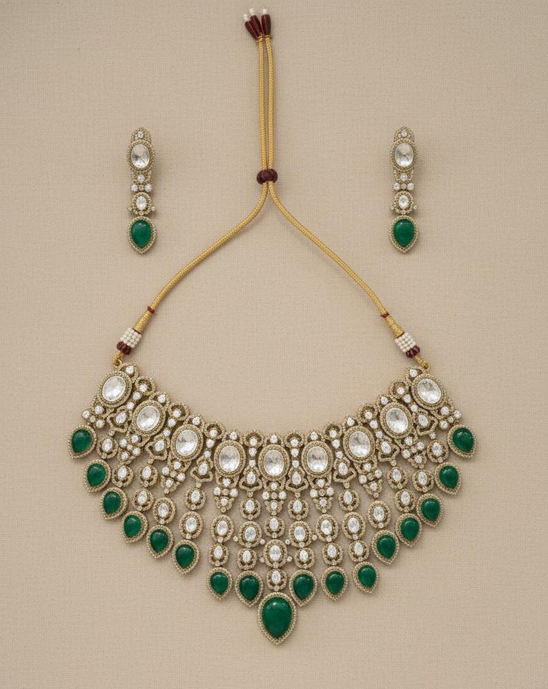 Op de afbeelding: Een goudkleurige ketting- en oorbellenset. De ketting heeft ovale en ronde heldere stenen met druppelvormige groene stenen. De oorbellen weerspiegelen het ontwerp met een enkele groene druppelsteen. De set wordt gepresenteerd op een neutrale achtergrond.