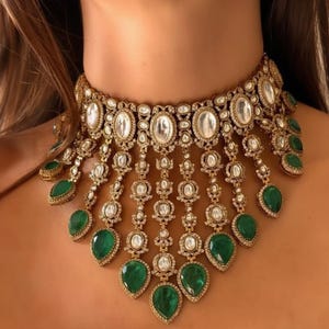 Op de afbeelding: Een sierlijke goudkleurige ketting met ovale en ronde heldere stenen, met bungelende, druppelvormige smaragdgroene stenen. De ketting is een choker-stijl, met ingewikkelde details en een vintage esthetiek.