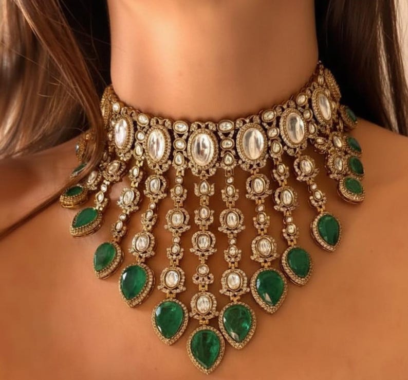 Op de afbeelding: Een sierlijke goudkleurige ketting met ovale en ronde heldere stenen, met bungelende, druppelvormige smaragdgroene stenen. De ketting is een choker-stijl, met ingewikkelde details en een vintage esthetiek.