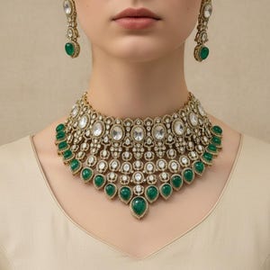 Op de afbeelding: Een goudkleurige ketting en bijpassende oorbellen set. De ketting heeft heldere, ovaal geslepen stenen en druppelvormige smaragdgroene stenen. De oorbellen hebben een vergelijkbaar ontwerp met een grote smaragdgroene steen.