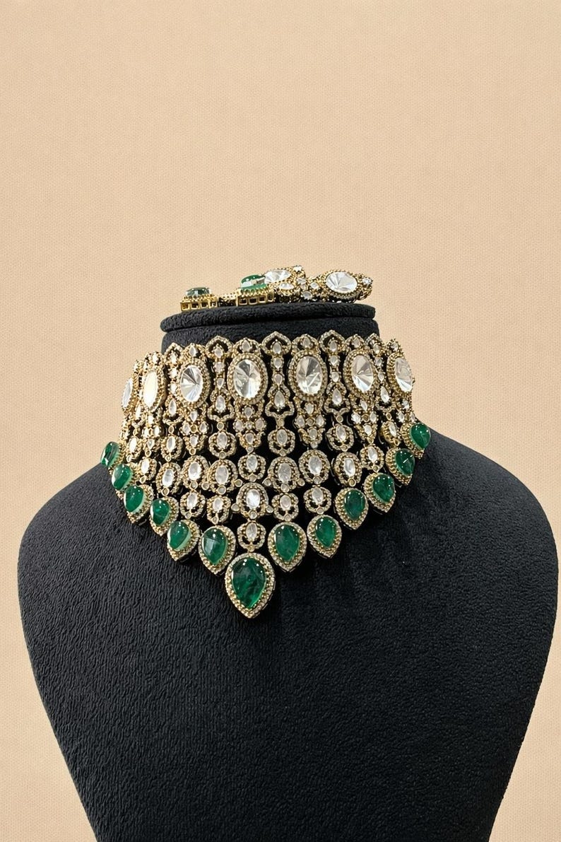 Op de afbeelding: Een sierlijke, goudkleurige ketting en bijpassende armband set. De ketting heeft grote, heldere ovaal geslepen stenen en kleinere heldere stenen, met druppelvormige smaragden. De sieraden worden getoond op een zwarte mannequin.
