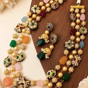 May include: A gold-coloured necklace and matching earrings set. The necklace features multiple strands of faux pearls, colourful gemstones, and decorative gold-coloured filigree elements with red, green, and clear accents. The earrings have a similar design.