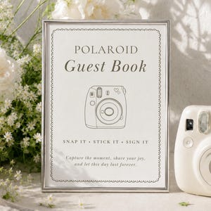 A silver-framed sign with white background and black text reading Polaroid Guest Book, Snap It Stick It Sign It, and Capture the moment, share your joy, and let this day last forever, featuring a line drawing of a camera.