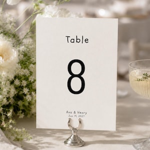 以下が含まれることがあります： Tableという文字と大きな黒い数字の8がシンプルなフォントで印刷された、長方形の白いテーブルナンバーカード。数字の下にはAva & Henryというテキストと2027年6月19日という日付が印刷されています。カードは銀色のスタンドに立てられています。