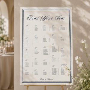 May include: A rectangular wedding seating chart sign with a cream background and a dark blue border. The text reads Find Your Seat at the top, followed by an alphabetical list of names and table numbers, and Elena & Maxwell at the bottom.