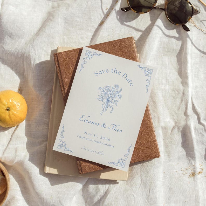 Op de afbeelding: Een trouwkaart met de tekst "Save the Date" in blauwe letters, met bloemenillustraties. De uitnodiging ligt op twee boeken, met een citroen en een zonnebril op de achtergrond. De datum is 17 mei 2026.