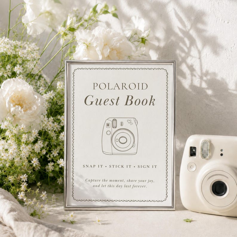 A silver-framed sign with white background and black text reading Polaroid Guest Book, Snap It Stick It Sign It, and Capture the moment, share your joy, and let this day last forever, featuring a line drawing of a camera.