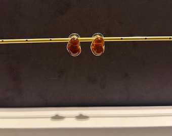 May include: A pair of amber-colored stud earrings with irregular, organic shapes. The earrings are set on gold-toned posts and displayed against a dark brown background. The earrings are a jewelry item.