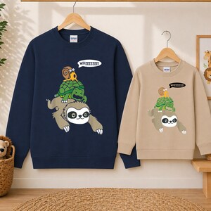 May include: Navy blue and tan sweatshirts with a whimsical graphic of a sloth carrying a turtle and snail, with the text "WHEEEEEEE!" in a speech bubble. The sweatshirts are hanging on a wooden rack.