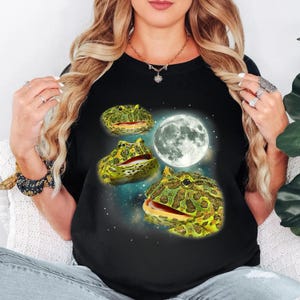 Può includere: T-shirt nero con un design stravagante di tre rane verdi e gialle con la bocca aperta, ambientate su un cielo stellato e una luna piena. Le rane hanno un aspetto strutturato e la luna è dettagliata con crateri.