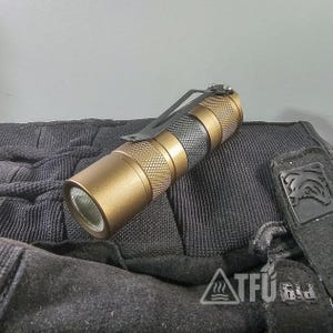 May include: A bronze-colored flashlight with a textured grip and a black pocket clip. The flashlight has a black and silver section in the middle. The image also shows a black glove with the text "ATFú" and a logo.