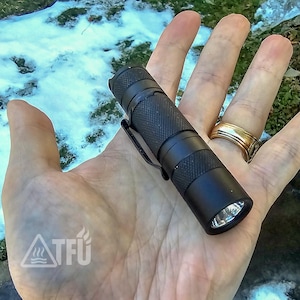 May include: A compact black metal flashlight featuring a textured knurled grip, a circular lens at the front, and a black metal pocket clip attached to the side. The cylindrical body has a matte finish and a small loop at the rear end.