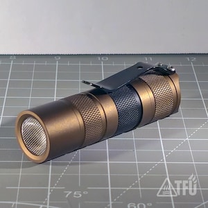 May include: A bronze-colored flashlight with a textured grip and a black pocket clip. The flashlight has a silver bezel around the light source and a black band in the middle. The flashlight is on a gray surface.
