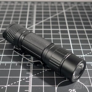May include: A black flashlight with a textured grip and a metal pocket clip. The flashlight has a bright LED light at the end and is lying on a black and gray grid surface.