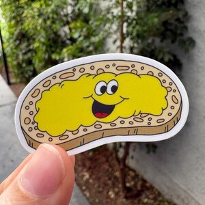 Könnte beinhalten: Ein Aufkleber mit einem Cartoon-Toastbrot mit einem lächelnden Gesicht. Der Toast ist braun mit einem gelben Belag, schwarzen Augen und einem roten Mund. Der Aufkleber wird von einer Hand gehalten.