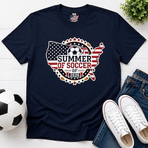 Könnte beinhalten: Ein dunkelblaues Kurzarm-T-Shirt mit einer Grafik der Landkarte der Vereinigten Staaten im Design der amerikanischen Flagge, einem Fußball und dem Text "SUMMER OF SOCCER OF 2026", umrandet von einem Kreis aus kleinen roten und weißen Sternen.