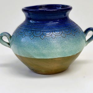 Könnte beinhalten: Eine handgefertigte Keramikvase mit zwei Henkeln. Die Vase hat einen Farbverlauf, der von einem tiefblauen Rand zu einer türkisfarbenen Mitte und einem braunen Boden übergeht. Dekorative Blumenmuster sind am oberen Rand eingeätzt.