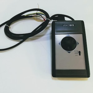 Può includere: Un'unità di controllo VIDEOSYS rettangolare nera e argento dotata di un joystick circolare centrale, un interruttore a tre posizioni con le diciture AUTO, MAN e OFF e un cavo elettrico nero collegato con fili interni multicolori esposti.