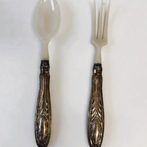 Może przedstawiać: Zestaw łyżki i widelca. Główki łyżki i widelca są w kremowobiałym kolorze. Uchwyty są ozdobne, z wykończeniem z metalu w kolorze srebrnym. Zestaw jest prezentowany na białym tle.