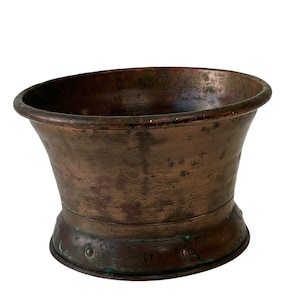 Könnte beinhalten: Ein verwitterter Kupfermörser mit einer breiten, ausgestellten Schüssel und einem abgerundeten Boden. Das Kupfer hat eine satte, gealterte Patina mit Anklängen von grüner Oxidation, die auf Alter und Gebrauch hindeuten. Das Design ist funktional und dekorativ.