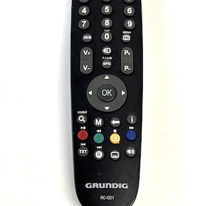 Puede incluir: Un mando a distancia negro Grundig RC-GD1 con numerosos botones, incluyendo números, volumen, canales y encendido. El mando tiene la palabra "MUTE" en verde y "POWER" en rojo. La marca "GRUNDIG" está impresa en la parte inferior.