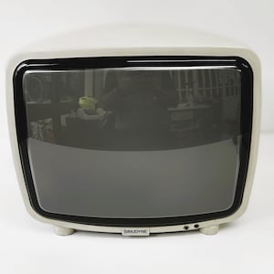 Könnte beinhalten: Ein cremefarbener Vintage-Fernseher mit schwarzem Bildschirm und Zierleisten. Der Fernseher hat eine abgerundete rechteckige Form und steht auf kleinen, abgerundeten Füßen. Der Markenname "SINUDYNE" ist auf der Vorderseite zu sehen. Der Bildschirm spiegelt die Umgebung wider.