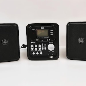 Puede incluir: Un sistema de audio negro Trevi que cuenta con una unidad central con pantalla digital, un gran dial de volumen plateado y varios botones de control, flanqueado por dos altavoces rectangulares negros a juego con rejillas y conos centrales.
