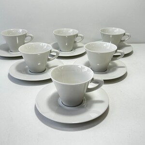 Peut inclure: Un ensemble de six tasses et soucoupes en céramique blanche disposées en formation triangulaire. Chaque tasse présente un corps lisse et conique avec une anse incurvée, tandis que les soucoupes circulaires servent de base à chaque tasse.