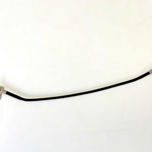 May include: A black flexible cable shutter release tool with a silver metal plunger mechanism at one end and a threaded metal connector at the opposite end. The device features a long, thin, curved black cord connecting the two metallic components.
