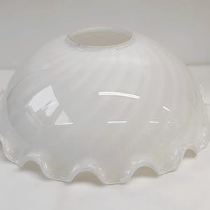 Może przedstawiać: Biały szklany klosz z falistą krawędzią. Klosz ma zaokrąglony kształt i wirujący wzór. Obrzeże ma muszlowy wzór. Klosz wykonany jest z błyszczącego, nieprzezroczystego szkła.