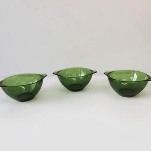 Op de afbeelding: Drie groene glazen kommen met een afgeronde vorm en kleine handvatten. De kommen zijn diep, transparant groen en staan op een wit oppervlak. De kommen zijn waarschijnlijk bedoeld om te serveren of te presenteren.