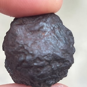 Può includere: Una roccia scura di forma irregolare con una superficie ruvida e strutturata. La roccia è tenuta tra due dita, mostrando la sua colorazione marrone scuro e nera. L'oggetto sembra essere una piccola pietra naturale.