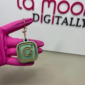 Könnte beinhalten: Ein mintgrüner Instagram-Logo-Schlüsselanhänger mit goldfarbenem Rand und Schlüsselring. Der Schlüsselanhänger wird von einer Person mit einem rosa Handschuh gehalten. Der Hintergrund zeigt den Text "la moo DIGITALLY" in Pink und Schwarz.