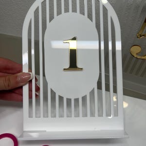 May include: A white acrylic table number sign featuring an arched top with vertical slats and a central oval cutout. A reflective gold-colored number one is attached to the center of the oval. The sign stands on a rectangular base.
