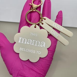 May include: A cream-coloured keychain with a gold clasp and ring. The keychain features a flower-shaped charm with the words "THIS mama BELONGS TO" in white script. Two rectangular tags are engraved with the names "OLIVIA" and "LIAM".
