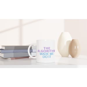 May include: White ceramic mug with the text "THE ALGORITHM MADE ME DO IT" in purple and blue gradient lettering. The mug is on a white surface with books and decorative vases in the background.