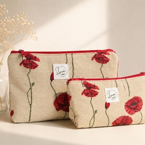 Può includere: Due pochette rettangolari con cerniera in tessuto beige con un motivo di papaveri rossi e steli verdi. Ogni pochette presenta un'etichetta quadrata bianca al centro con la parola Love stampata in nero e una chiusura a zip rossa.