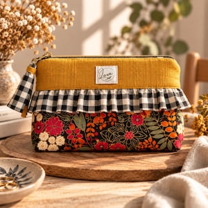Pode incluir: Uma bolsa retangular com fecho de correr, apresentando uma faixa superior amarelo-mostarda com uma etiqueta branca com a palavra Love, um folho axadrezado preto e branco e uma secção inferior com fundo preto e padrões florais coloridos.