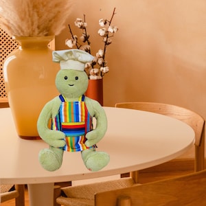 May include: A plush green turtle wearing a white chef's hat and a colorful striped apron. The turtle is sitting on a round, light-colored table. A large yellow vase and a vase with cotton stems are in the background.