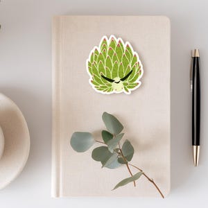 May include: A green sticker with a cartoon-style design of a plant with a face, placed on a beige notebook. The plant has a spiky, layered appearance with red accents. A black pen and a sprig of greenery are also visible.
