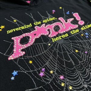 Peut inclure: Sweat &agrave; capuche noir avec un motif "POOK!" rose orn&eacute; de strass, une toile d'araign&eacute;e et le texte "nevermind the spider, heres the slime". Le motif comprend des &eacute;toiles et des pierres pr&eacute;cieuses.