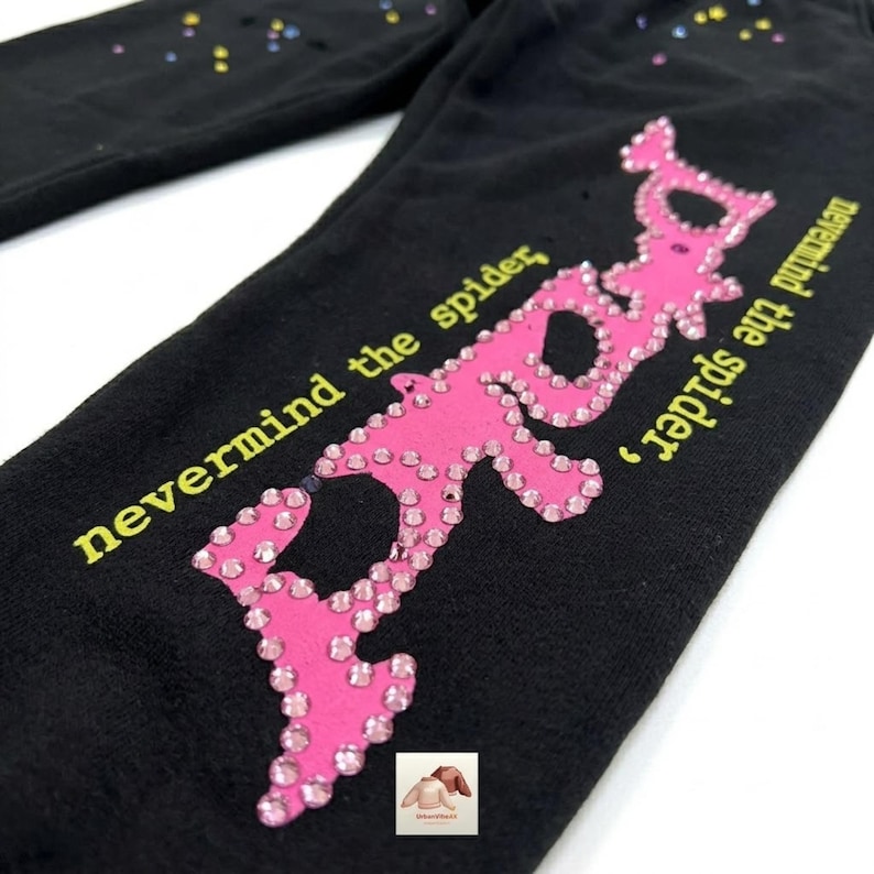 Peut inclure: Pantalon de surv&ecirc;tement noir avec un motif d'araign&eacute;e rose orn&eacute; de strass et le texte "nevermind the spider" en jaune. Le motif de l'araign&eacute;e est soulign&eacute; de strass. De petits strass color&eacute;s sont dispers&eacute;s sur le pantalon.