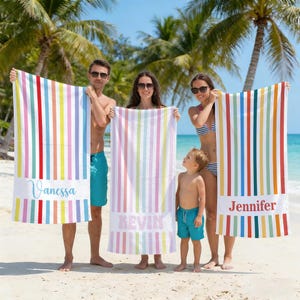 May include: Three colorful beach towels with vertical stripes in red, blue, yellow, and green. Each towel has a white band with a name printed on it: "Vanessa", "Kevin", and "Jennifer". The towels are held by people on a sunny beach.