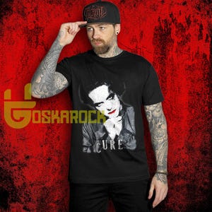 May include: Black t-shirt featuring a grayscale image of a person with red lipstick and the word "CURE" below. The person is wearing a black baseball cap with white text. The background is red.