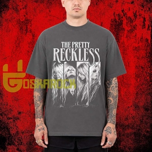 May include: A charcoal gray short-sleeve t-shirt featuring the white text THE PRETTY RECKLESS above four vertical panels displaying black and white photographic portraits of a person with long hair, set against a red textured background.