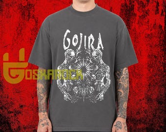 May include: A charcoal gray short-sleeve t-shirt featuring a white graphic print on the chest. The design displays the text GOJIRA above a detailed illustration of two skeletal figures flanking a central sunburst and intricate organic patterns.